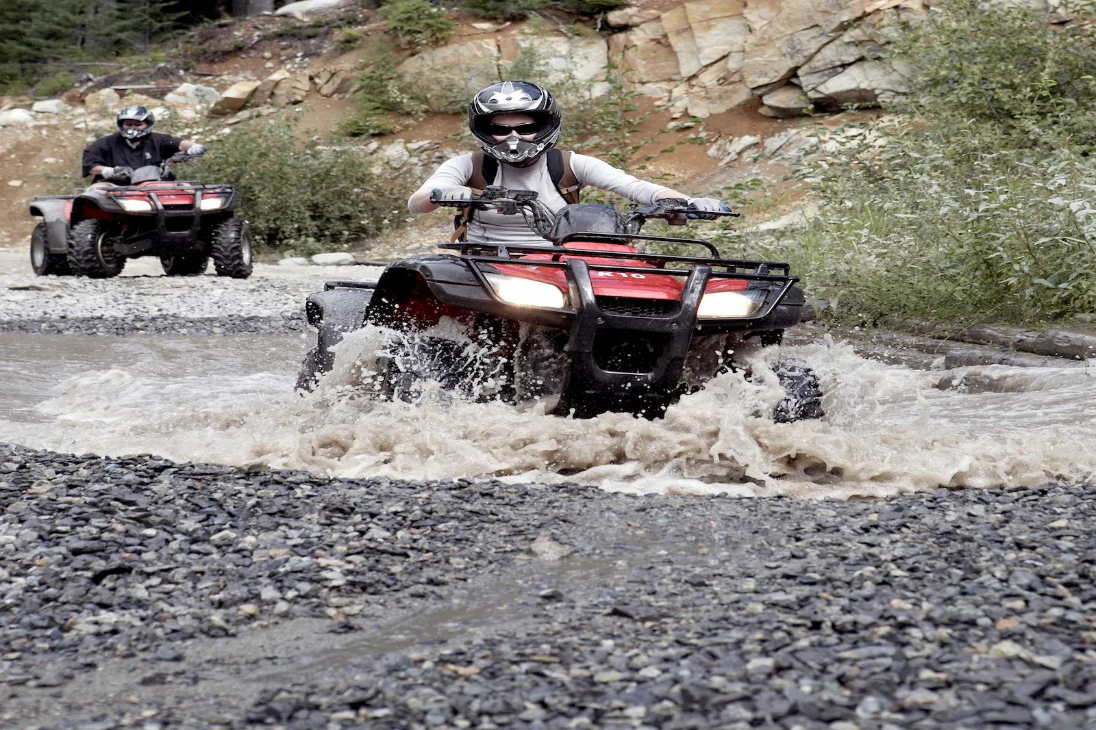 ATV riders crossing a rocky stream on an off-road trail, representing recreational vehicle insurance coverage for ATVs and UTVs.