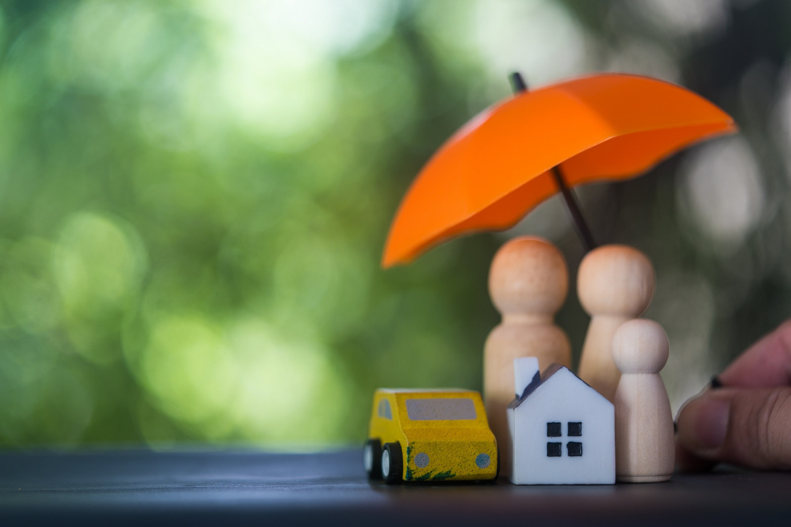 Wooden toy house, car, and family under an umbrella, representing umbrella insurance protection for assets and liability.
