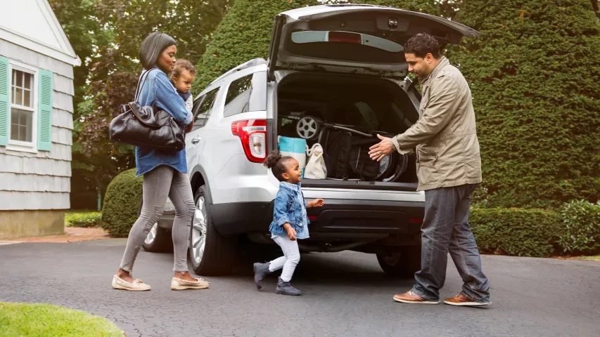 Family with two young children preparing to travel, loading belongings into a car, representing auto insurance protection.
